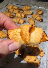 Load image into Gallery viewer, A hand holds a small, golden-brown Apricot Rugelach with fruit filling. More Apricot Rugelach pastries rest on a parchment-lined baking sheet in the background, making them an ideal festive holiday treat.